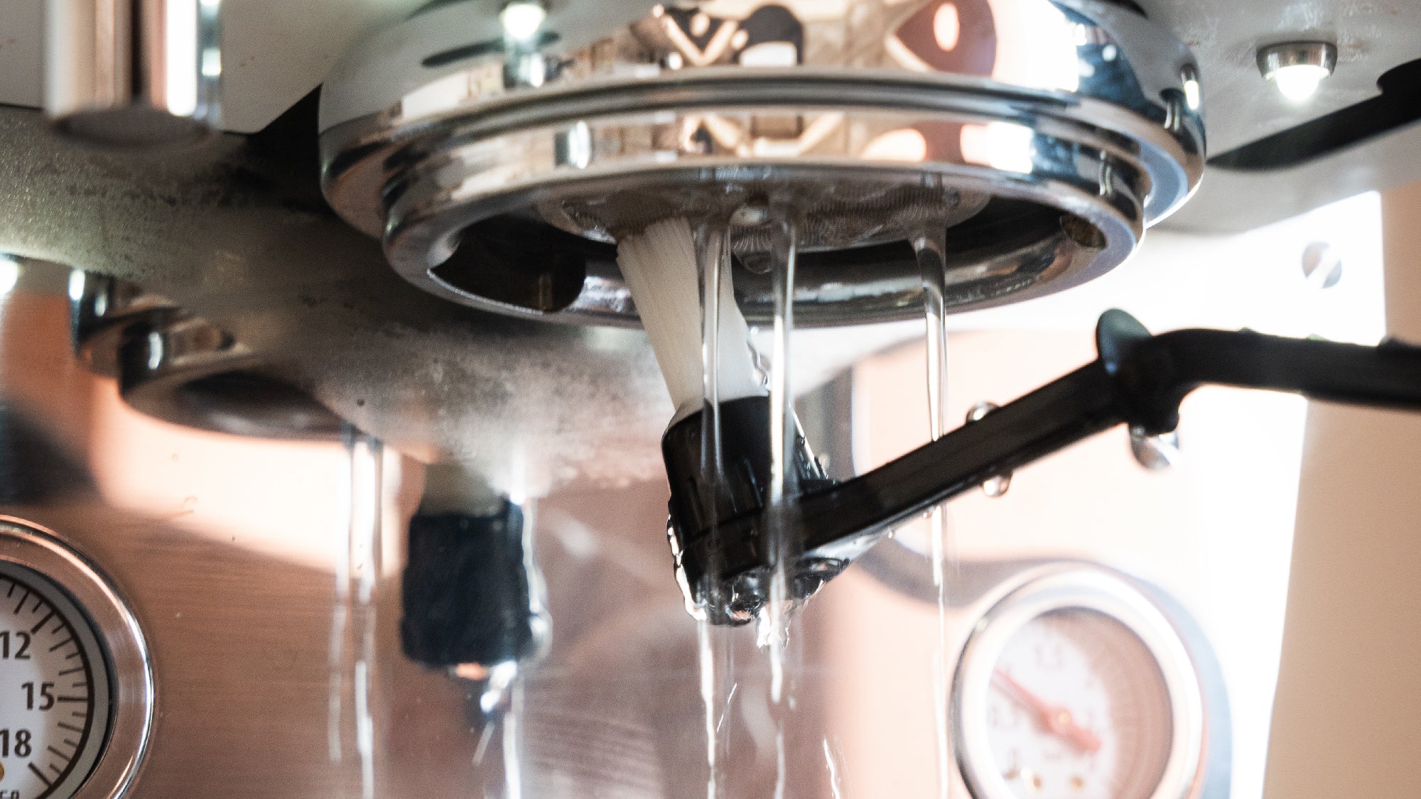 Een borstel reinigt de zetgroep van een espressomachine terwijl er heet water uitstroomt.